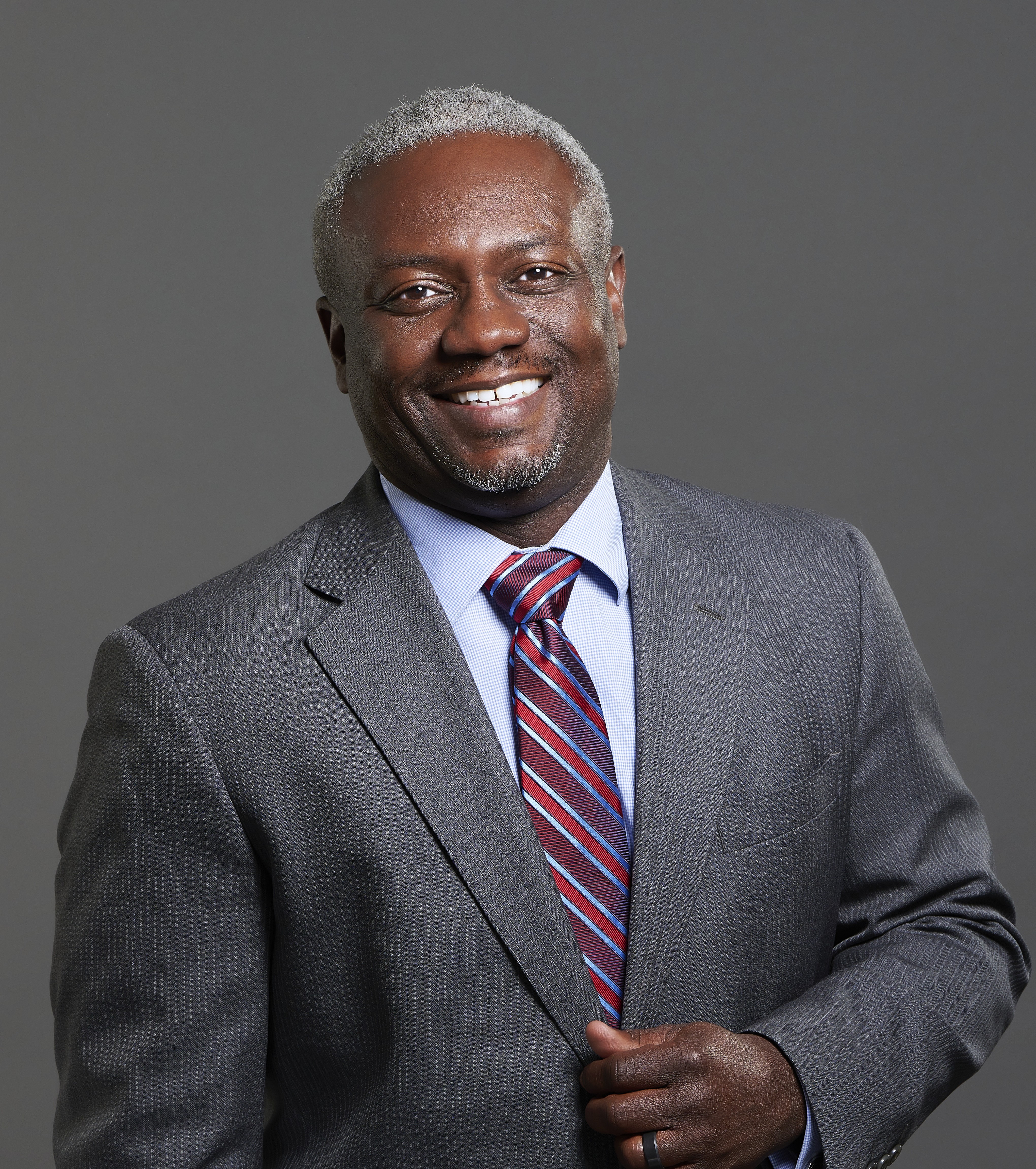 Black man with short grey hair smiling in a grey suit and striped red tie